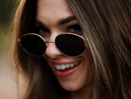 Fotografia di Drimage Creative studio, realizzata dalla fotografa Simona Turano. Primo piano di una donna sorridente con occhiali da sole rotondi neri e lunghi capelli castani, sullo sfondo sfocato di un paesaggio esterno.