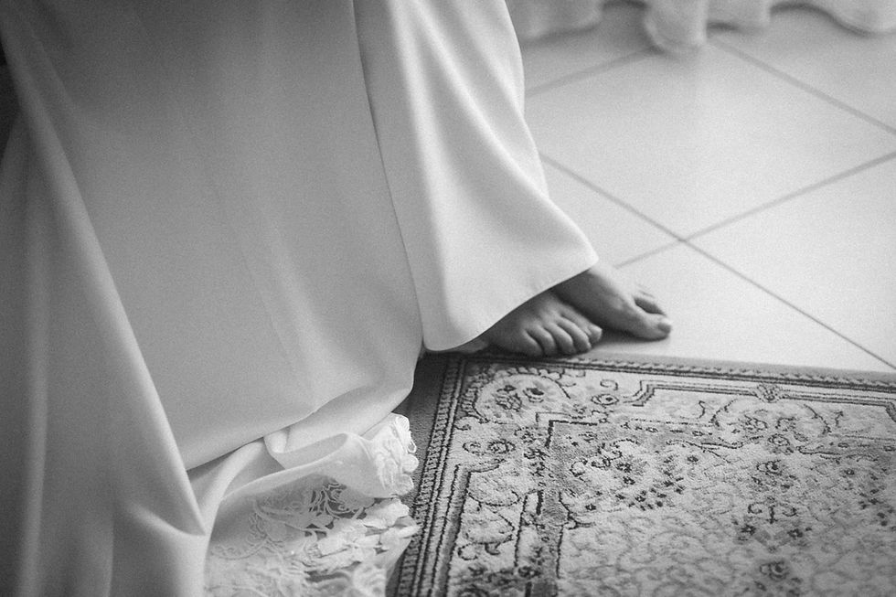 Fotografia di Drimage Creative studio di Simona Turano Fotografa. Primo piano di piedi nudi che fanno capolino da un abito bianco svolazzante su un tappeto decorato, creando un'atmosfera serena e intima. Ambientazione monocromatica.