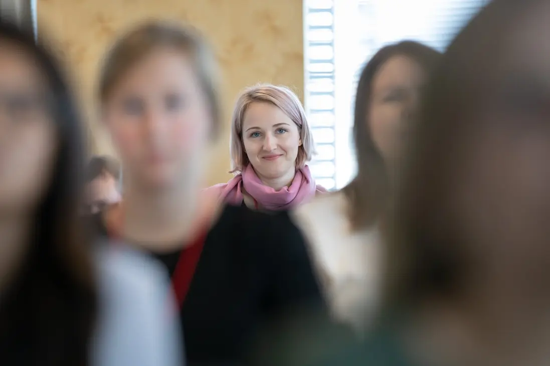 A business portrait of a woman in a pink scarf, sharply focused among a blurred crowd, conveying leadership and individuality.