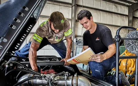 Two workers inspecting and referencing a manual while servicing a large vehicle engine.