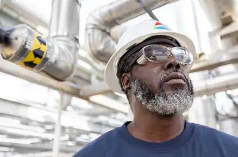 A professional business portrait of an industrial worker in a hard hat and safety goggles at a piping facility.