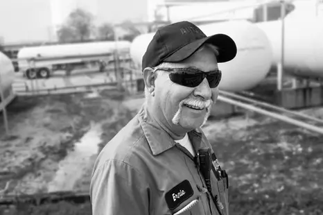 A business portrait of a smiling industrial worker wearing sunglasses and a cap, standing near large outdoor storage tanks.