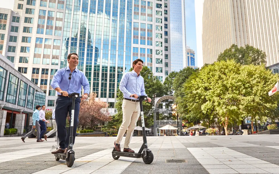 Two men ride scooters through a sunny downtown plaza in lifestyle product photography.