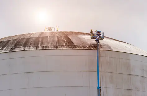 Worker power-washing the roof of a large industrial tank from a boom lift under bright sunlight.