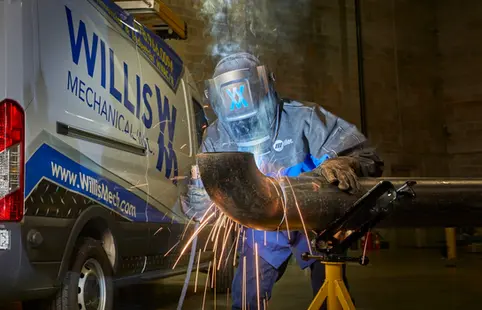 Welder wearing a protective helmet working on a pipe near a company van, with sparks flying.