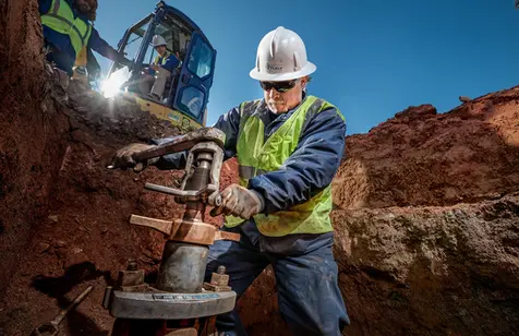 Worker operating valve installation tool in a trench, with crew and excavator in the background.