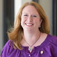 Corporate headshot photography of a red-haired woman in a purple blouse and pendant necklace, smiling outdoors.