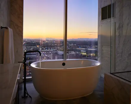 Freestanding bathtub in luxury hotel bathroom overlooking city skyline at sunset.