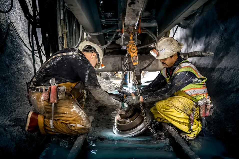 Two construction workers adjusting or installing a part on machinery underground.