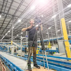 Photographer Gregory Campbell on a platform films a modern warehouse interior using a camera on a tripod.