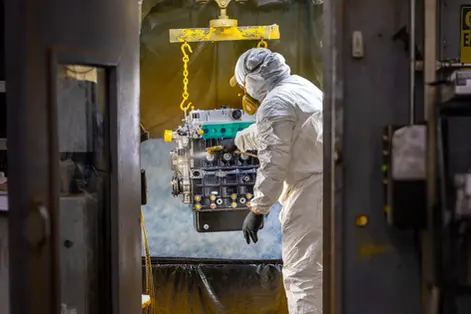 Worker in a full protective suit and mask spray-painting a suspended engine block.
