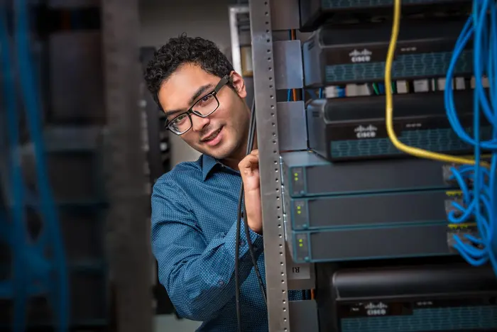 A professional business portrait of an IT technician smiling while managing cables in a server room.
