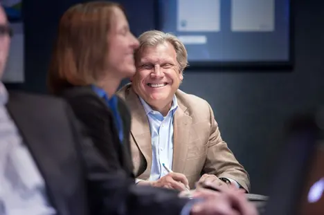 A professional business portrait of a man in a tan blazer laughing in a conference room during a meeting.