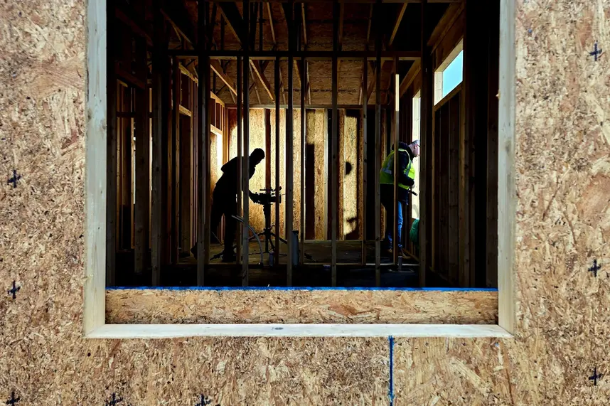 Workers silhouetted inside a wood-framed building under construction, viewed through an open window frame.