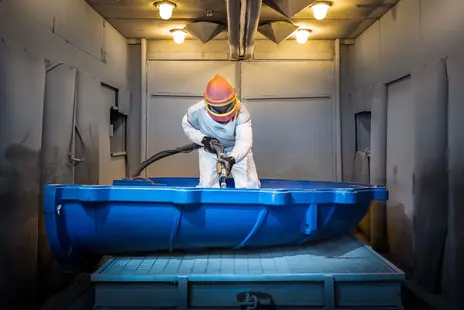 Worker in protective suit and helmet using a hose or blaster on a large blue part in a booth.