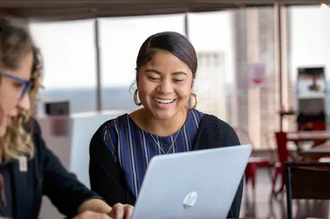 A woman smiles while working on a laptop in a company café, reflecting warmth and focus in corporate lifestyle.