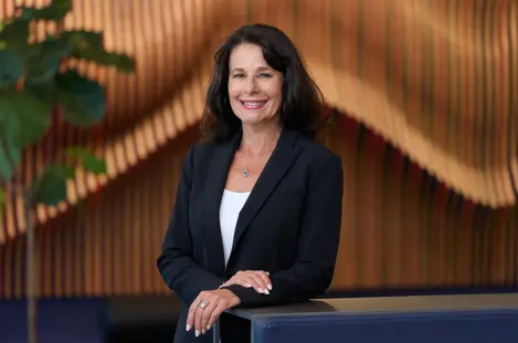 A professional business portrait of a woman in a blazer, smiling confidently in a modern office lobby.