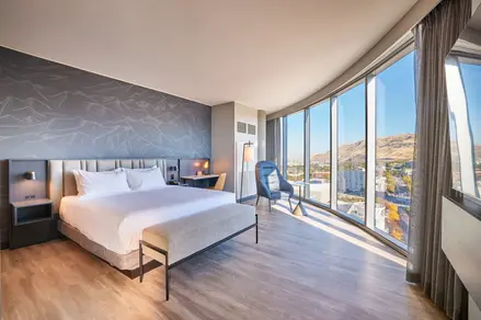 Hyatt Salt Lake City corner guestroom with bed, reading chair, and panoramic mountain views through curved glass.