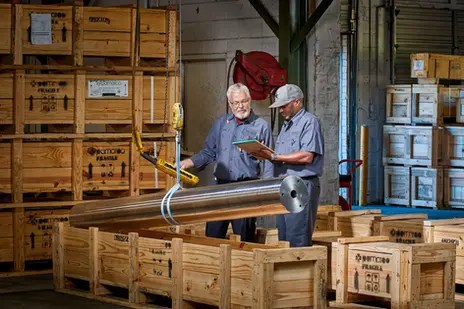 Two employees review paperwork while inspecting a large metal roller in a crate-filled warehouse.