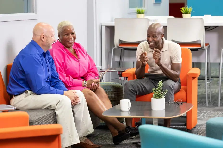 Three coworkers talk in a lounge with plants and soft seating, capturing relaxed collaboration in corporate lifestyle.