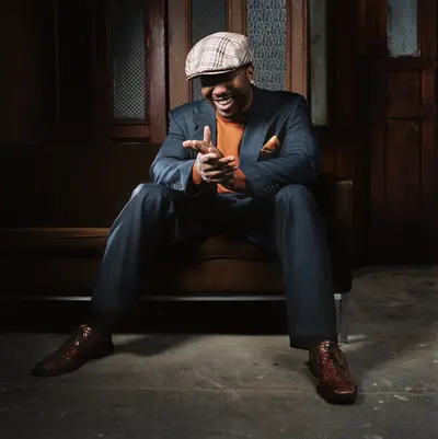 A professional business portrait of a stylish artist in a suit and cap, seated confidently in a warmly lit interior.