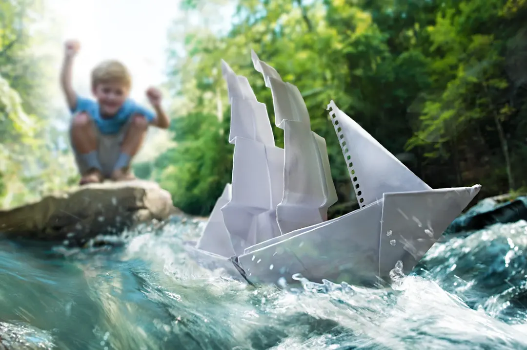 Paper sailboat floats in stream with excited child in lifestyle product photography.