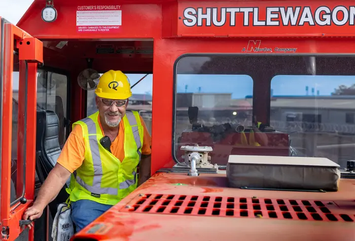 A business portrait of a rail yard equipment operator smiling while exiting a red shuttle wagon in work gear.