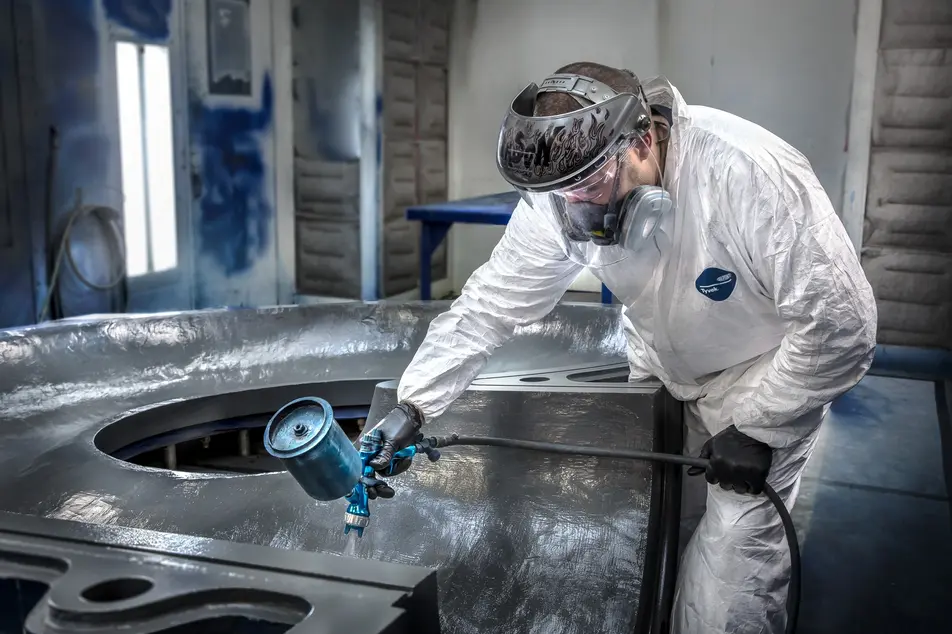 Worker in protective gear spray-painting a large metal industrial part in a paint booth.
