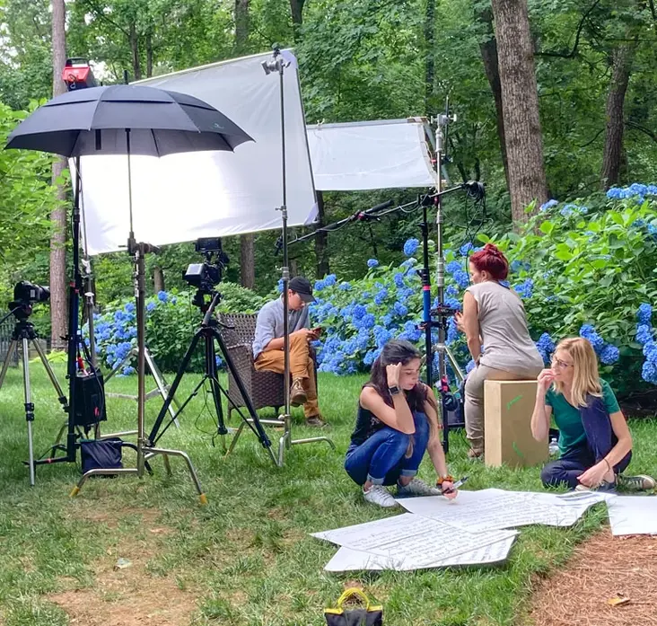 Film crew reviews a script on set with cameras and lighting near blooming hydrangeas in a wooded area.