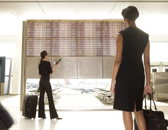 Women check flight info at airport in business attire for lifestyle product photography.