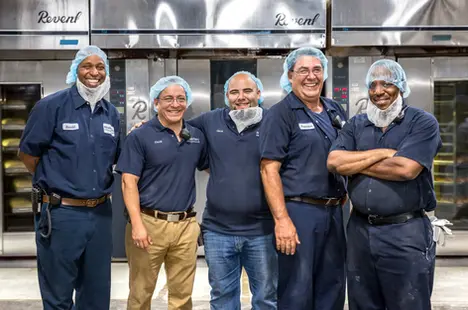 A professional business portrait of five bakery employees in uniform, smiling and standing side by side in front of commercial ovens.