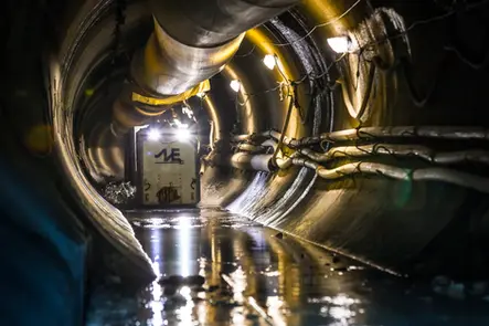 Underground tunnel with reflective wet floor, utility lights, and mining vehicle approaching.