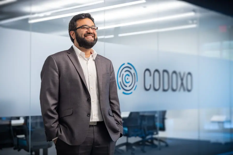 A professional business portrait of an executive standing confidently in an office with the Codoxo logo in the background.