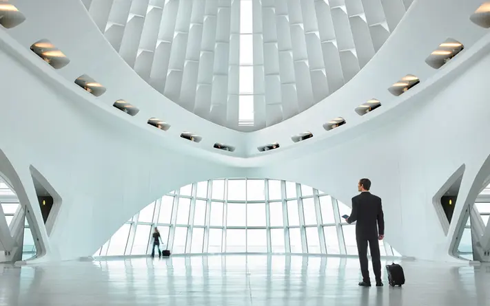 Interior architectural photography of a sleek modern terminal with curved white walls, geometric ceiling panels, and travelers passing through the space. Keywords: interior architectural photography, commercial interior photography, modern terminal.