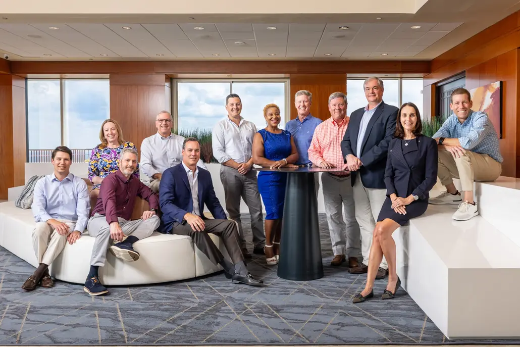A professional business portrait of an executive leadership team posing together in a modern office lounge.