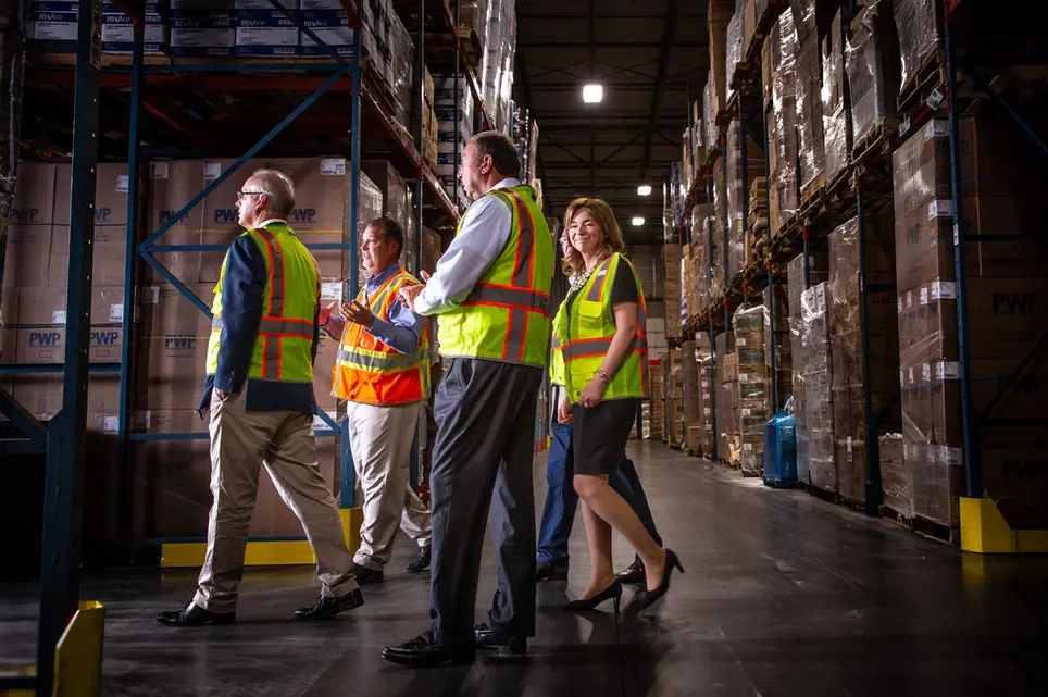Team in safety vests tours a warehouse, highlighting operations and leadership in corporate lifestyle industries.