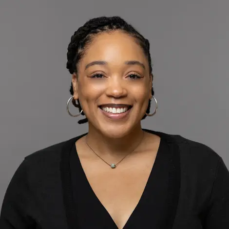Corporate headshot photography of a smiling woman in a black top with hoop earrings and a small necklace, against a gray backdrop.