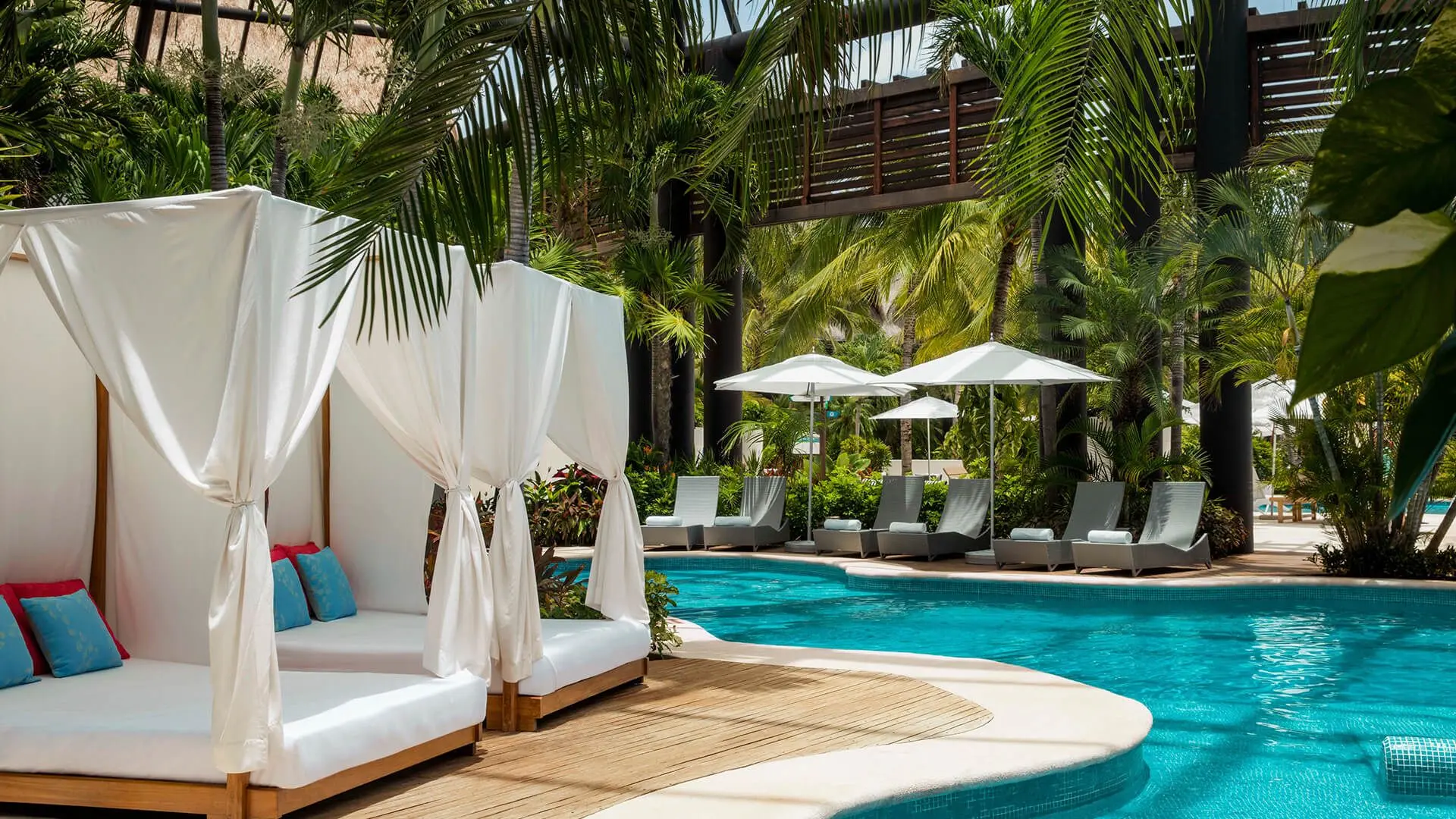 vibrant blue pool waters next to cabanas draped in white linens under green palm trees and vegetation 