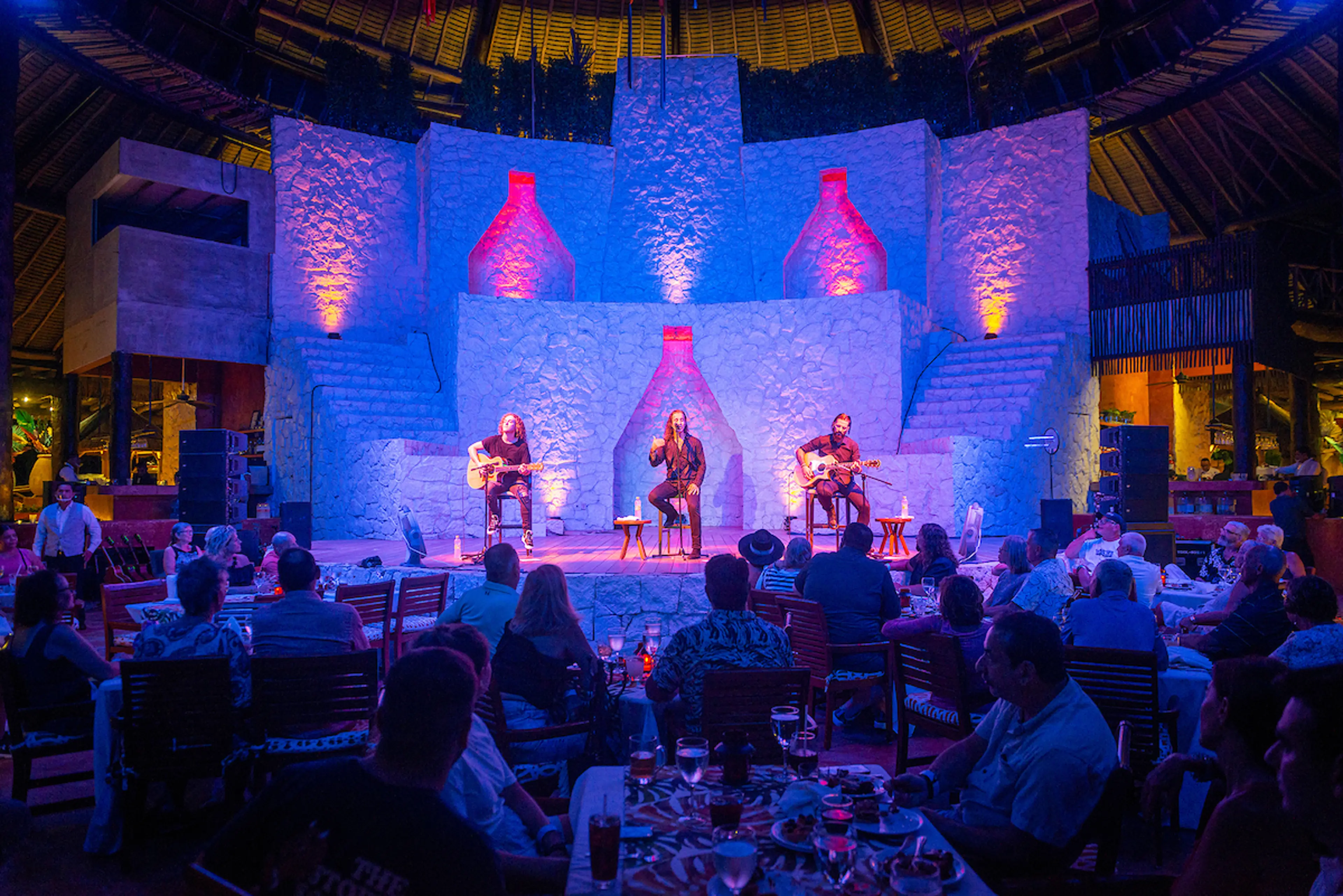 3 performers with 2 guitars in front of a live audience with illuminated backdrop 