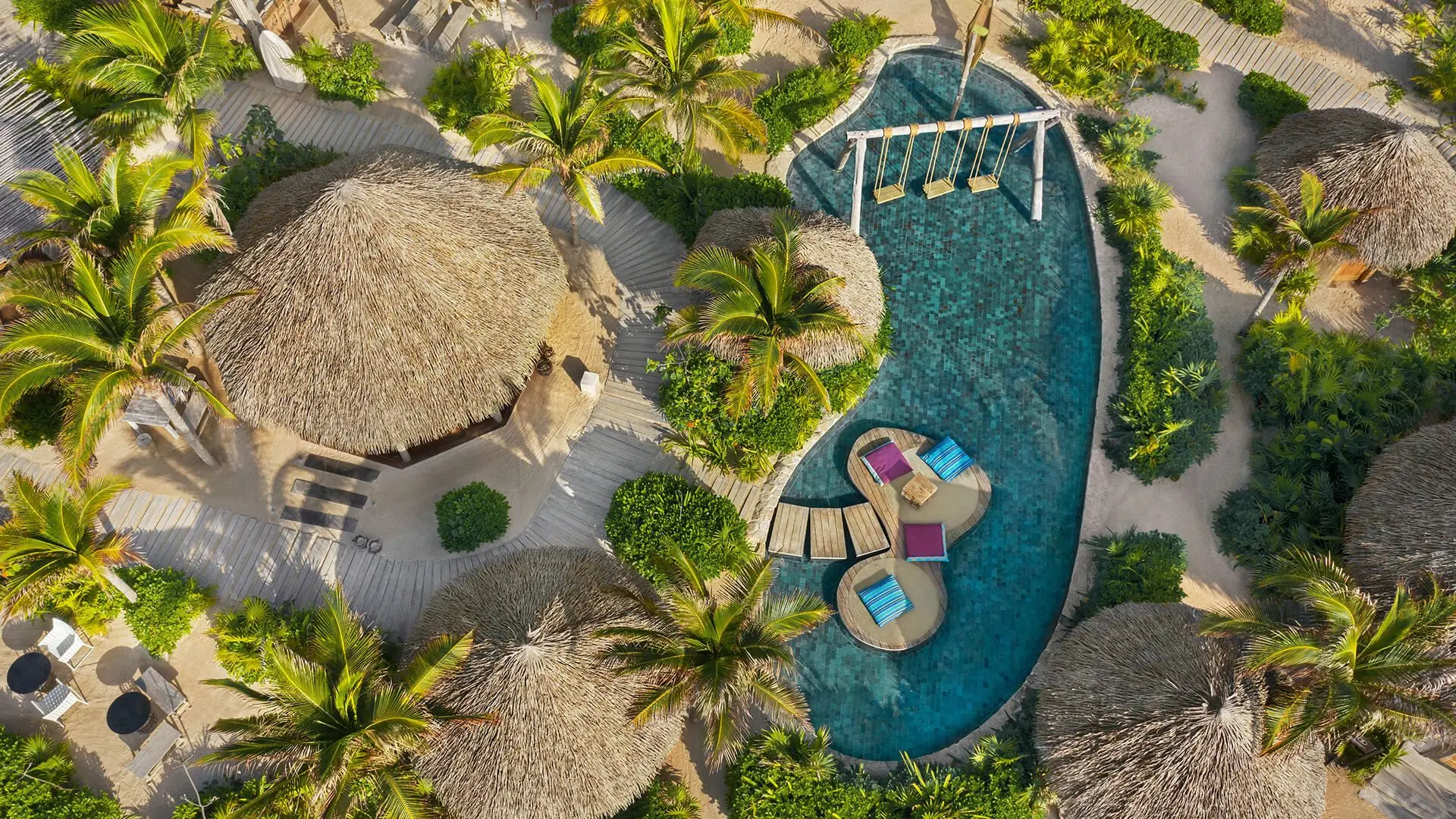 vibrant palm trees and cabanas next to a blue pool with large pillow chairs and swings 