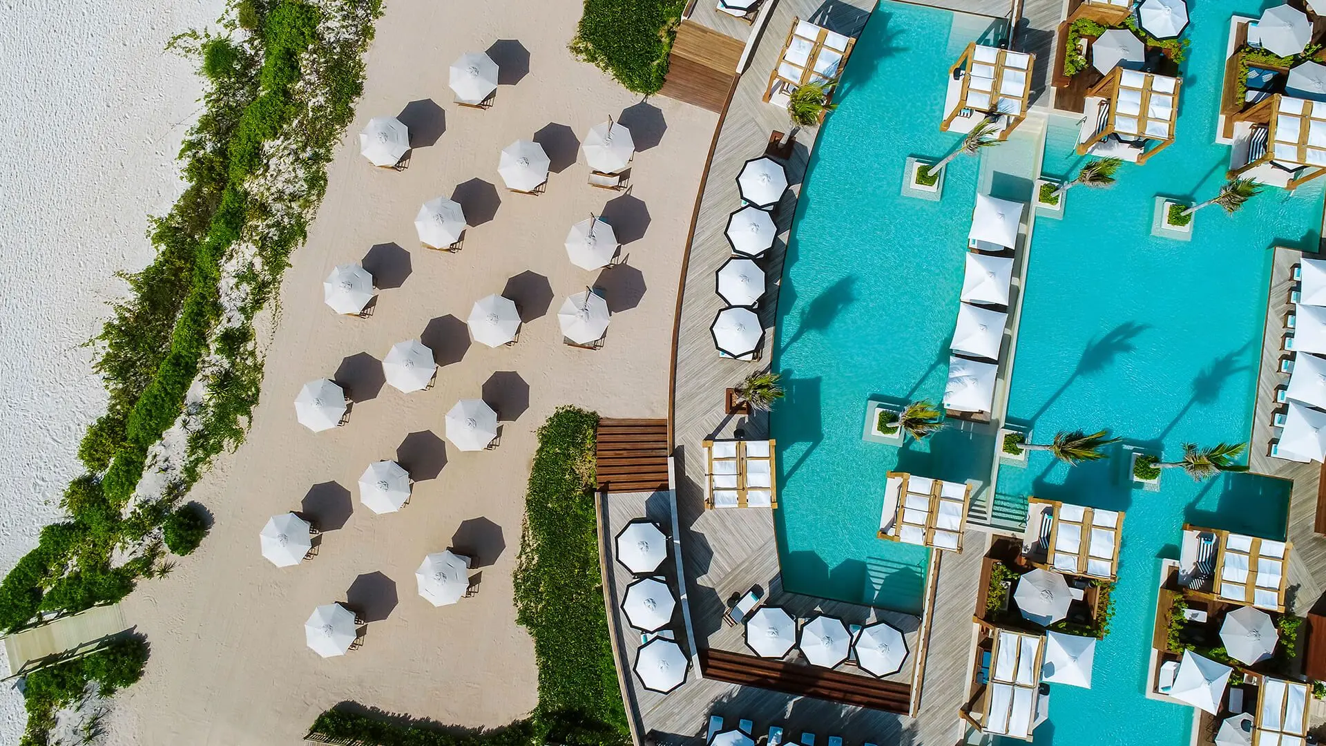 aerial view looking down at white beach and pool umbrellas with a shadow cast to the side of it next to white sand beach and a pool