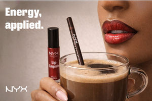 Close-up of a person holding a glass cup of coffee with NYX lip products placed alongside and inside the cup, with bold text overlay reading “Energy, applied.”
