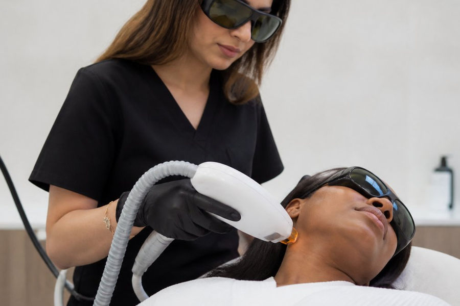 Professional dermatologist performing laser hair removal treatment on patient with melanated skin in modern clinical setting, showing laser handpiece positioned on skin with protective eyewear visible