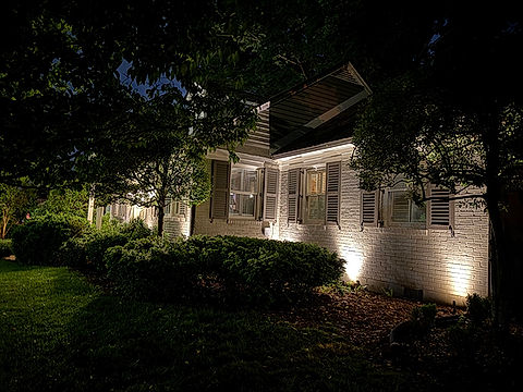 White house with lights at night. Trees and bushes in front.