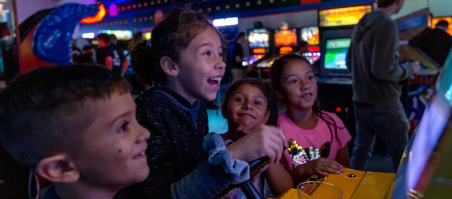 a group of kids watching a girl play an arcade game smiling