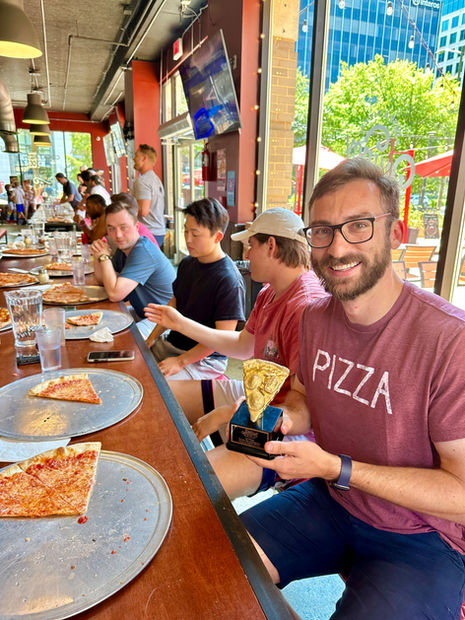 Man in 'PIZZA' shirt holding a slice, smiling with friends at a counter