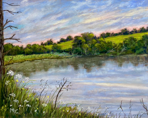 A small lake with a green hills at the back foreground countryside flowers and a pink and blue sunset