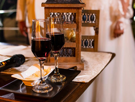 foto de detalhe da cerimônia-mesa com duas taças de vinho e casal de noivos ao fundo, desfocados