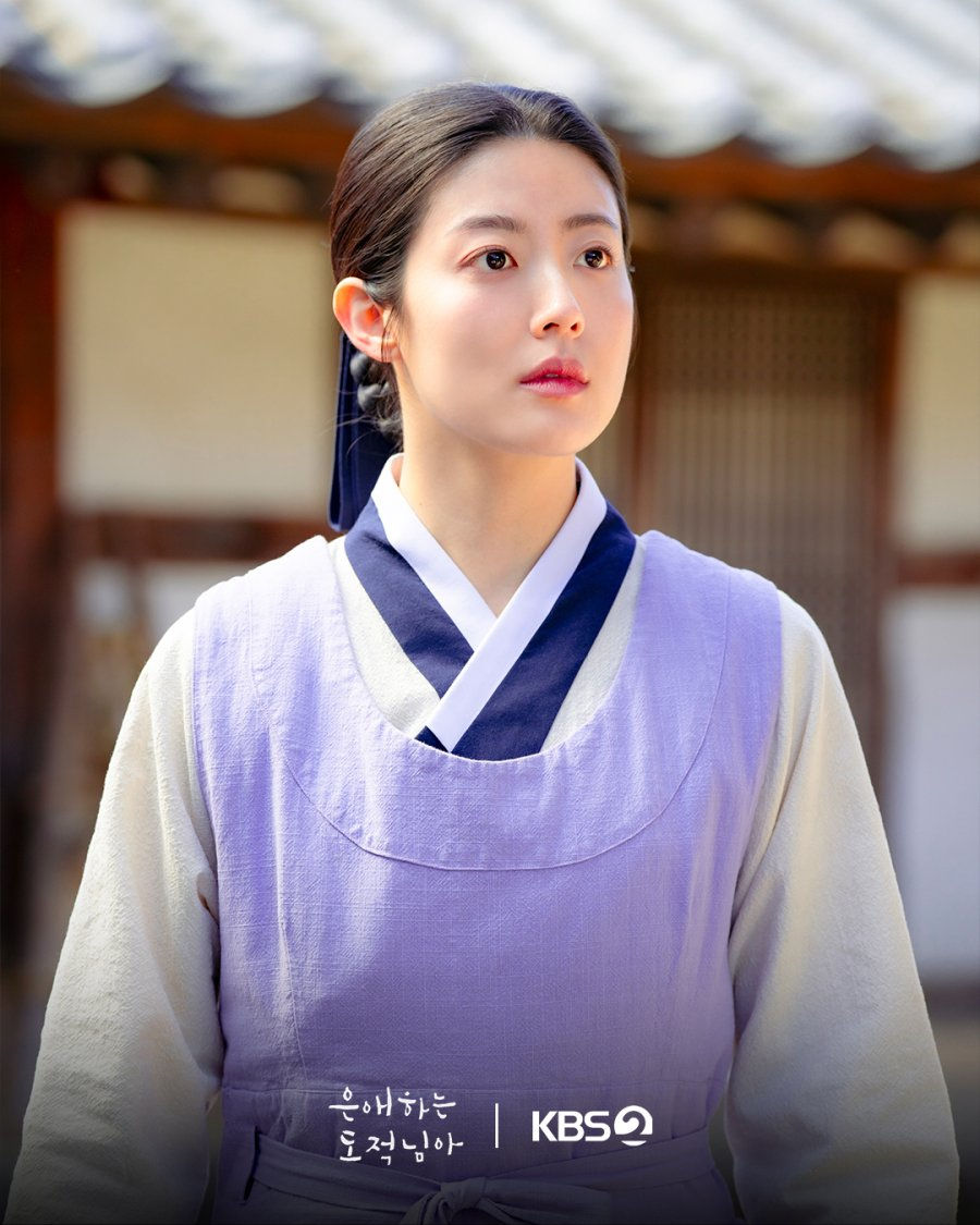 Young woman in lavender hanbok stands in a traditional courtyard, looking upward with a calm, pensive expression; To My Beloved Thief