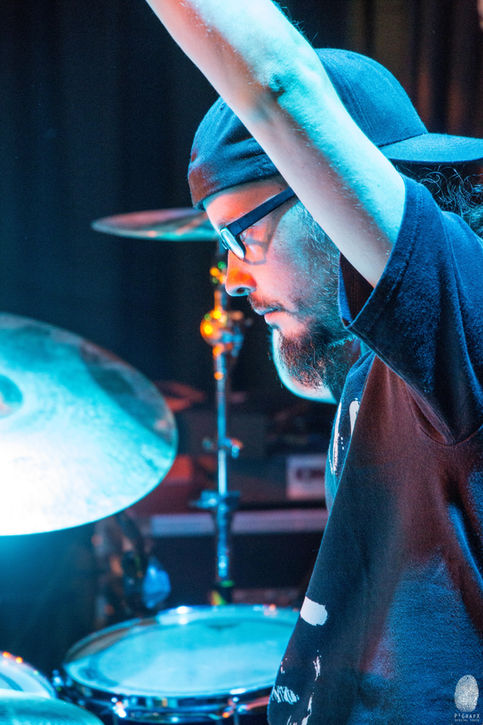 a man wearing glasses and a hat stands in front of a drum set